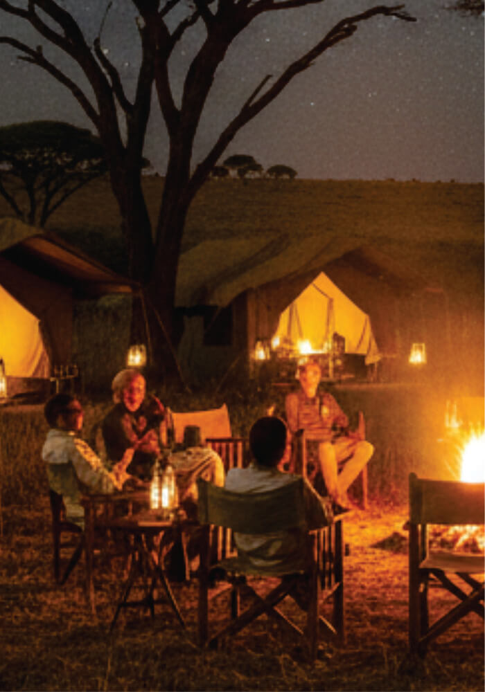 People sitting around a campfire at night in a safari setting with tents and trees.