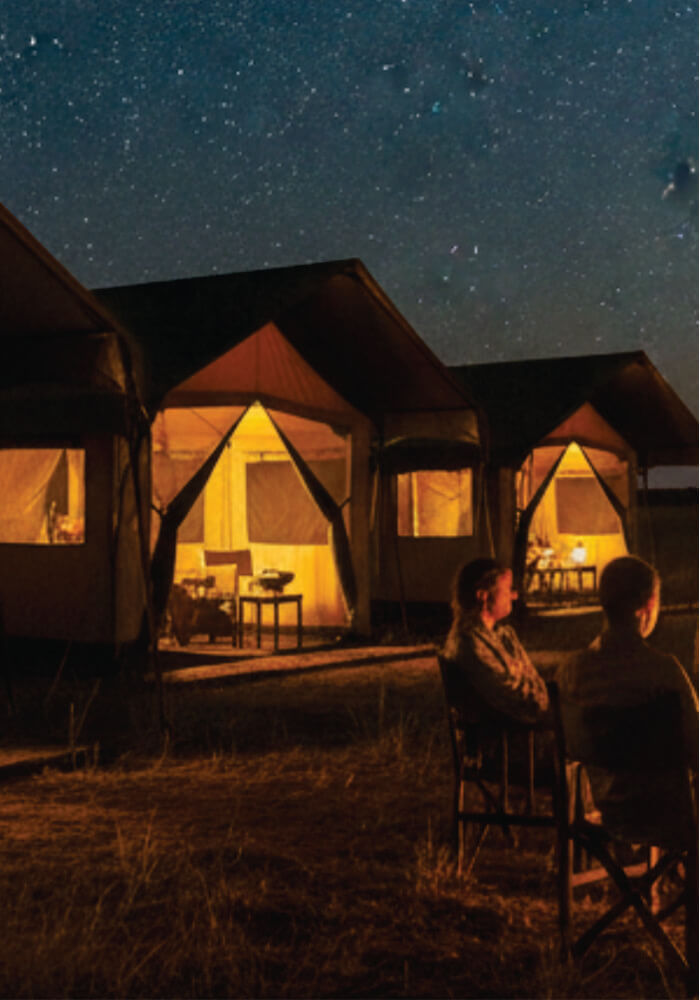 Two people sitting in front of lit safari tents under a starry night sky.