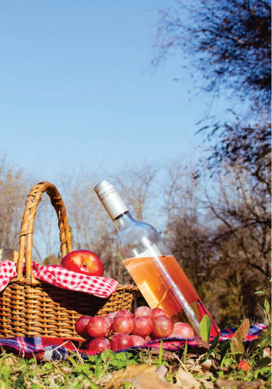 Wicker picnic basket with a bottle of rosé, apples, and grapes on a grassy outdoor setting.