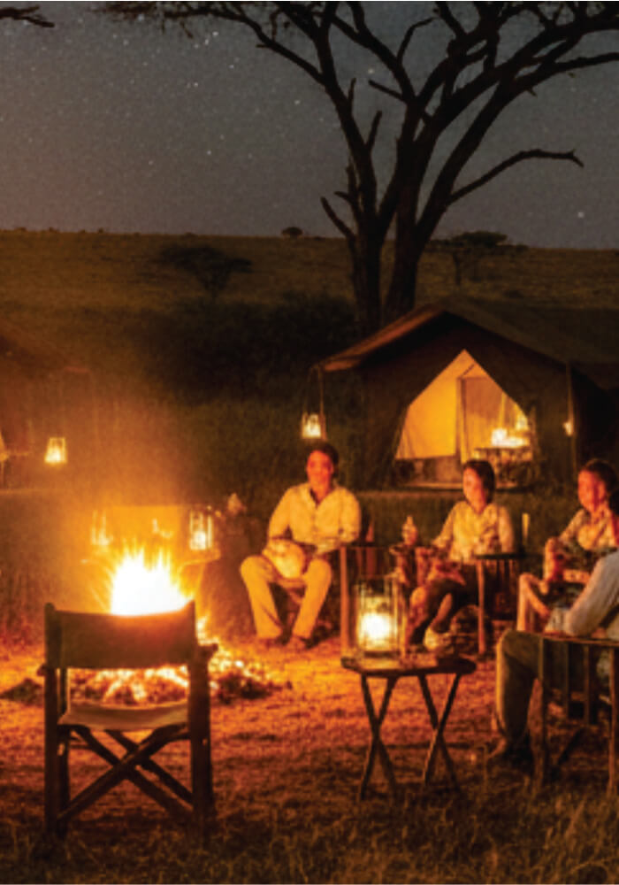 People sitting around a campfire at night in a safari setting with a tent in the background.