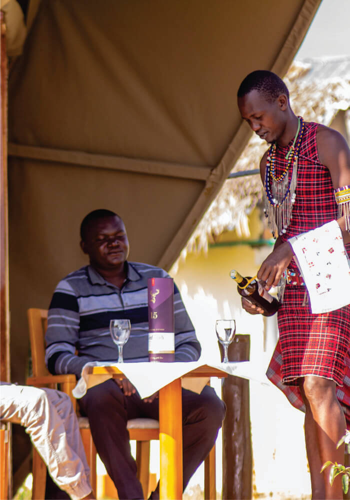 waiter putting on red Masai Shuka and serving a whole bottle of wine to a table of guest on budget luxury accommodation in Masai Mara Hotels